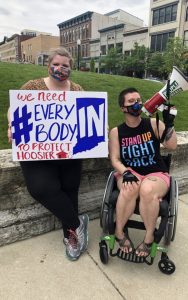 two people, one standing holding a sign that says "we need everybody IN to protect Hoosier homes" and the other in a wheelchair and holding a megaphone